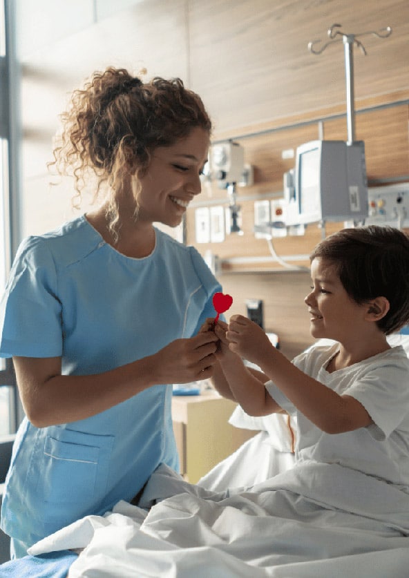 Kinderkrankenpflegerin in hellblauer Arbeitskleidung gibt Kind einen Herzlutscher im Krankenbett